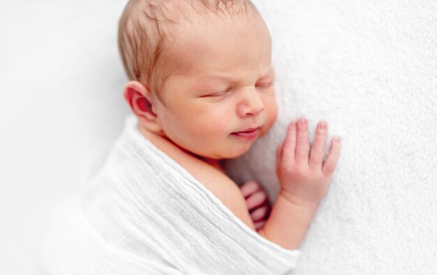 白い柔らかい布に包まれて眠っている新生児の男の子のクローズアップの肖像画。美しい幼児のプロの写真撮影。小さな子供が彼の頬の下で昼寝と手を握っています