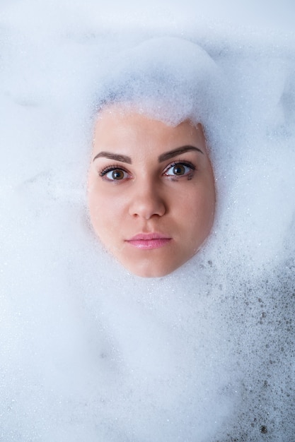Фото Портрет крупного плана девушки в ванне и белой пене вокруг ее лица. разные эмоции, гримасы, женская улыбка
