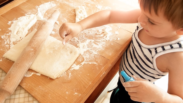 麺棒で小麦生地を圧延し、特別なプラスチック カッターでクッキーを切る愛らしい 3 歳の幼児の少年のクローズ アップの肖像画