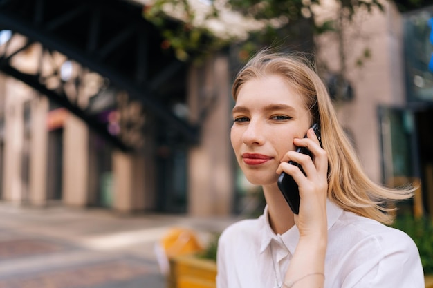 写真 電話で話していると街の通りに立っているカメラを見て笑顔の幸せな金髪の若い女性のポートレート、クローズ アップ