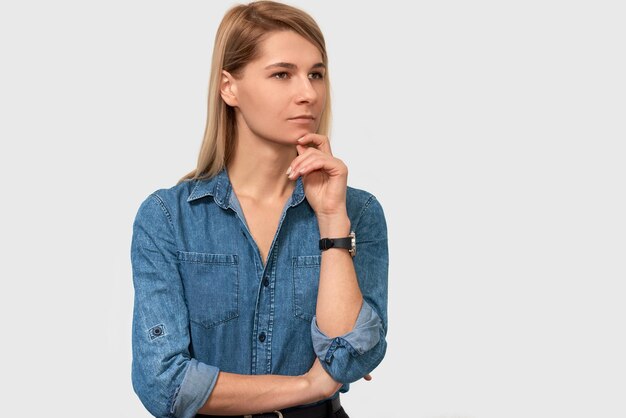 Closeup portrait of pensive beautiful young woman in denim shirt with finger folded frowning her face and looking to the one side on white studio background Thoughtful female thinking about something