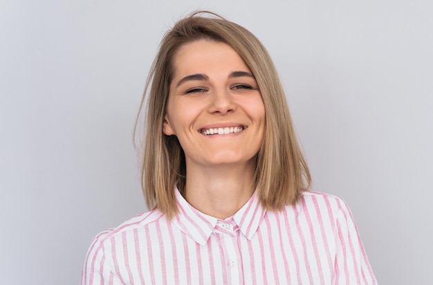 Closeup portrait of positive smiling blonde female with attractive look and beautiful green eyes wearing pink shirt posing against white wall in studio Happy woman showing positive emotions