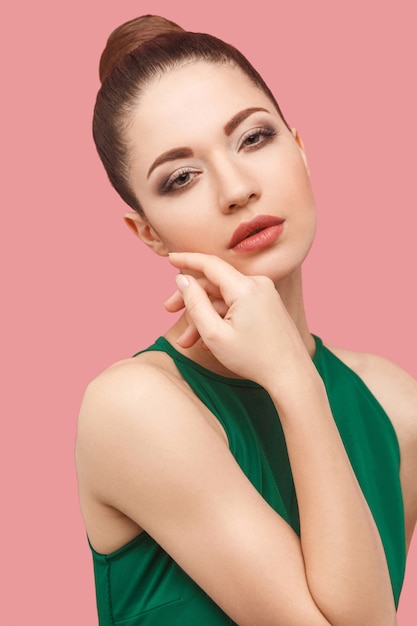 Closeup portrait of serious calm beautiful young woman with bun hairstyle and makeup in green dress standing touching her face and looking at camera. indoor studio shot, isolated on pink background.