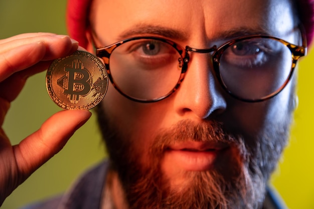 Foto ritratto del primo piano del ragazzo serio dei pantaloni a vita bassa con gli occhiali che mostrano moneta d'oro bitcoin criptovaluta, denaro virtuale elettronico. studio indoor girato isolato su sfondo colorato con luce al neon.