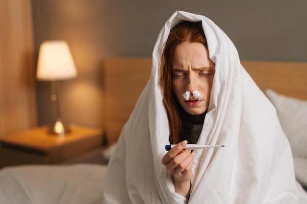 Closeup portrait of sick sad woman suffering from runny nose disease with paper tissues into nose measuring temperature with thermometer