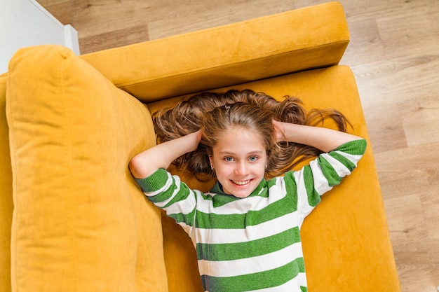 A closeup portrait of a smiling beautiful little blonde girl lying on a yellow sofa top view