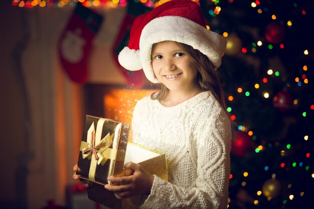 Ritratto del primo piano della ragazza sorridente in berretto di babbo natale in posa con scatola regalo incandescente