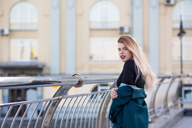 Closeup portrait of stylish blonde woman wearing green coat holding onto the rail. Space for text