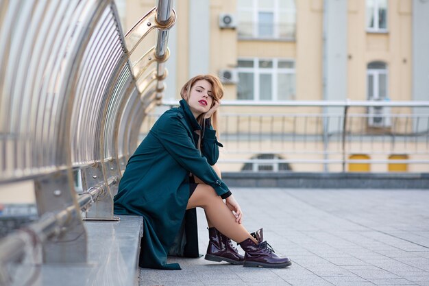 Closeup portrait of stylish blonde woman wearing green coat sitting near the handrail. Space for text