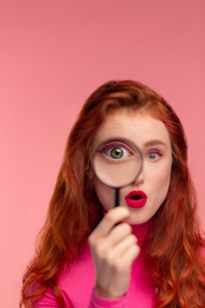 Closeup portrait of surprised redhead girl looking at camera through magnifying glass