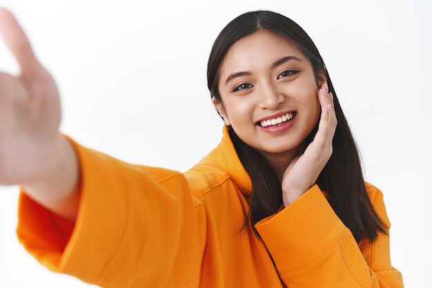 Closeup portrait tender pretty asian girl in orange hoodie touching pretty clean face and smiling taking selfie on mobile phone stretch hand to hold camera make cute expression white background