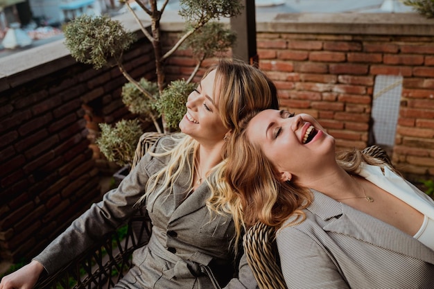Closeup portrait of two female friends in strict gray suits laughing while sitting on wicker chairs