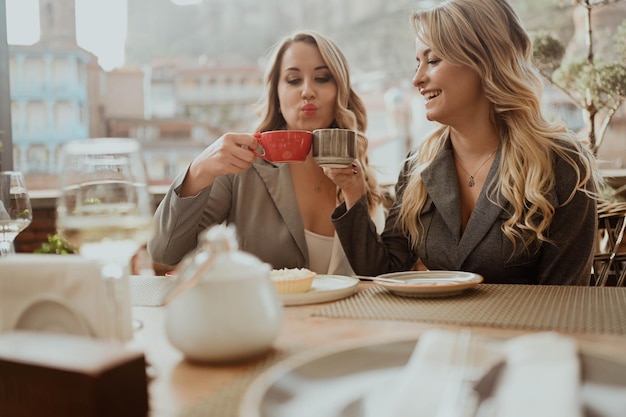 Closeup portrait of two female friends in strict suits laughing drinking coffee and wine on the