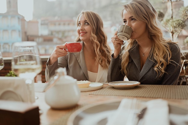 Closeup portrait of two female friends in strict suits laughing drinking coffee and wine on the