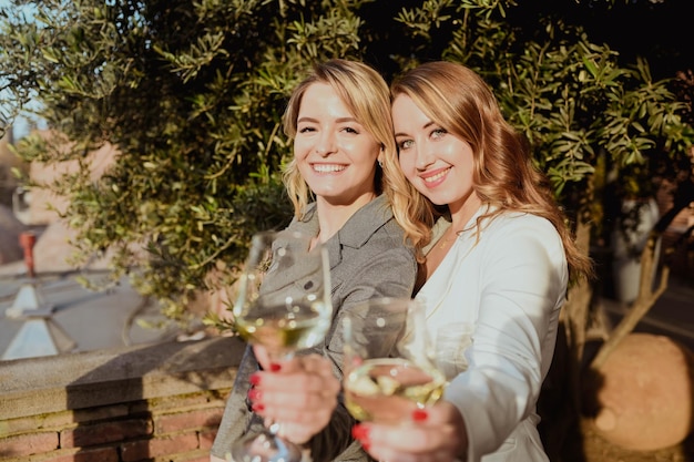 Ritratto del primo piano di due amiche in abiti rigorosi che ridono bevendo vino sulla terrazza all'esterno