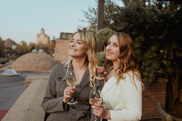 Closeup portrait of two female friends in strict suits laughing drinking wine on the terrace outside