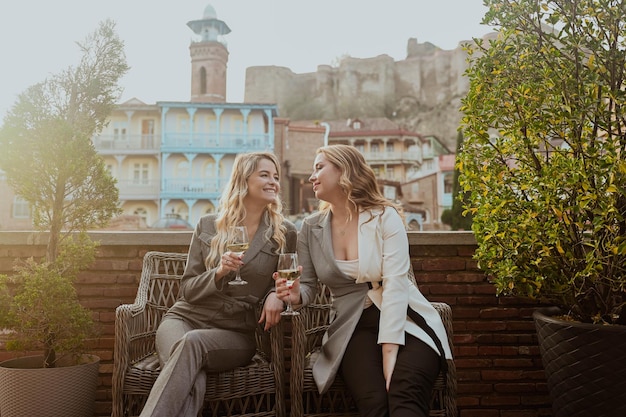 Ritratto del primo piano di due amiche in abiti rigorosi che ridono bevendo vino sulla terrazza all'esterno