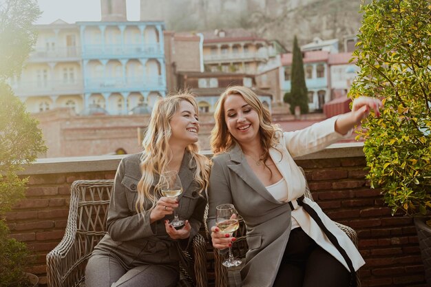 Closeup portrait of two female friends in strict suits laughing drinking wine on the terrace outside