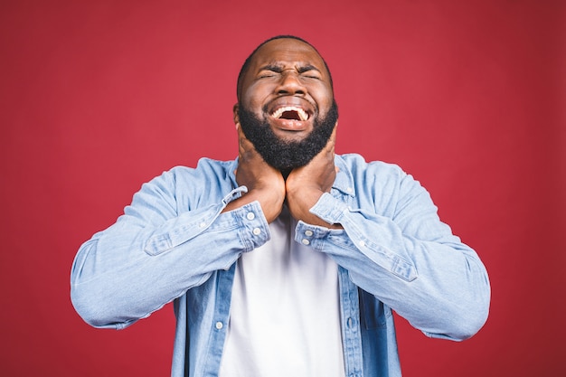 Closeup portrait upset stressed young african american man squeezing his head, going nuts, screaming, losing his mind, isolated red wall background.