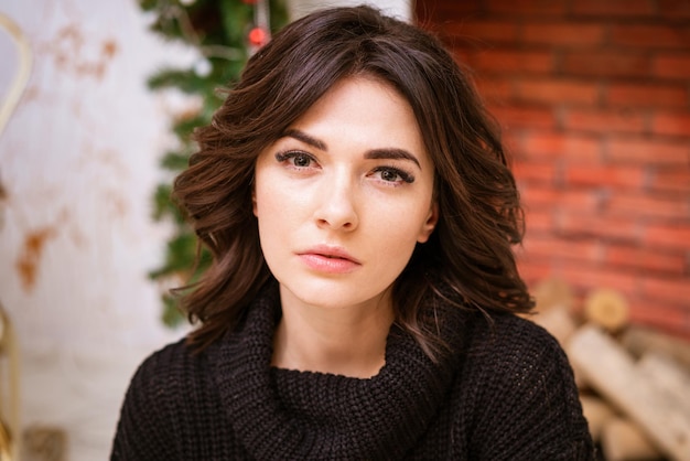 Closeup portrait of woman looking at camera against background of fireplace with christmas decor bea...