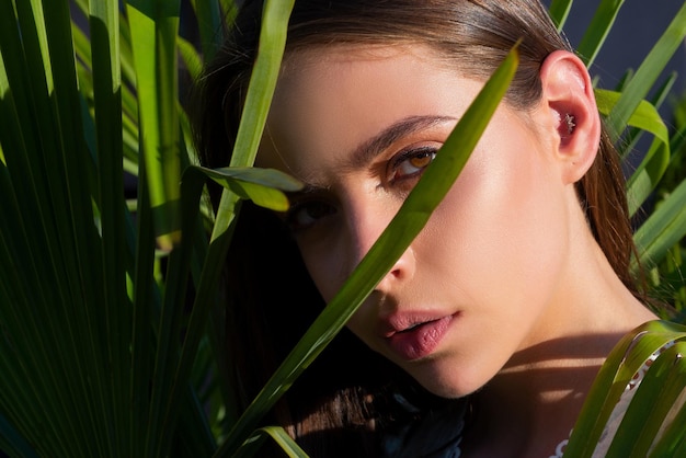 Closeup portrait of young and beautiful woman with perfect smooth tanned skin in tropical leaves con