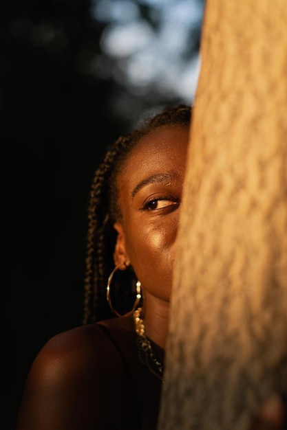 Closeup portrait of a young black woman hiding half of her face behind a tree in the park