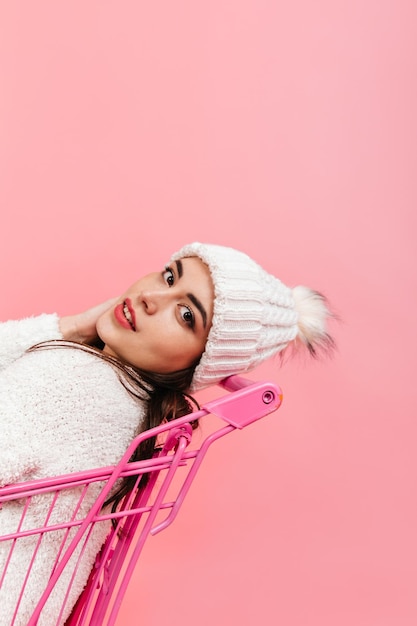 Closeup portrait of young darkhaired girl in white hat and sweater sitting in bright supermarket trolley on pink background