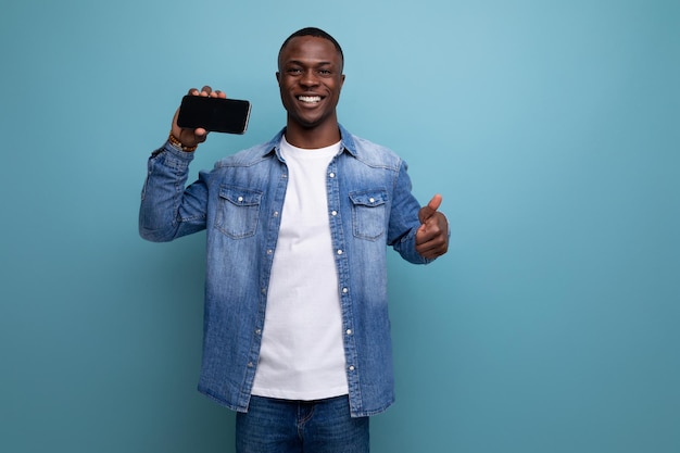 Photo closeup portrait of young handsome african guy in denim jacket holding smartphone with mockup on