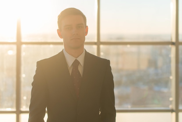 Closeup portrait of a young handsome businessman looking seriously at camera standing in light office over the large window background