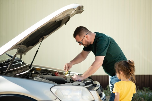 Closeup portrait, young man with his daughter having trouble with his broken auto, opening hood trying to fix engine, isolated green trees outside background. Car won't start, dead battery