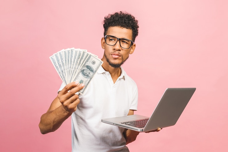 Photo closeup portrait, young successful african american business man making money from internet, holding cash in hand, laptop in another