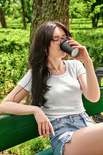 Closeup portret jonge mooie vrouw koffie drinken uit zwarte papieren beker zittend op een bankje in het park