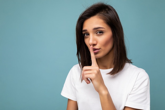 Closeup portret van jonge aantrekkelijke brunette vrouw met oprechte emoties dragen casual wit t-shirt geïsoleerd op blauwe muur met kopie ruimte en stil gebaar tonen.