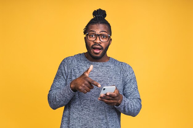 Closeup portret van knappe jonge Afro-Amerikaanse zwarte man, geschokt, verrast, wijd open mond, gek door wat hij op zijn mobiele telefoon ziet, geïsoleerd op gele achtergrond.