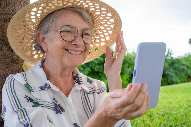 Closeup portret van lachende aantrekkelijke senior vrouw met hoed en bril zitten in openbaar park met behulp van mobiele telefoon kaukasische dame genieten van vrije tijd met mobiele telefoon