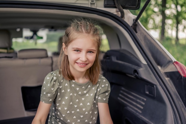 Closeup portret van schattig preteen meisje met mooi kapsel zittend in de kofferbak en glimlachend kijkend naar de camera. Gelukkig kind in het voertuig in de natuur tijdens de zomervakantie