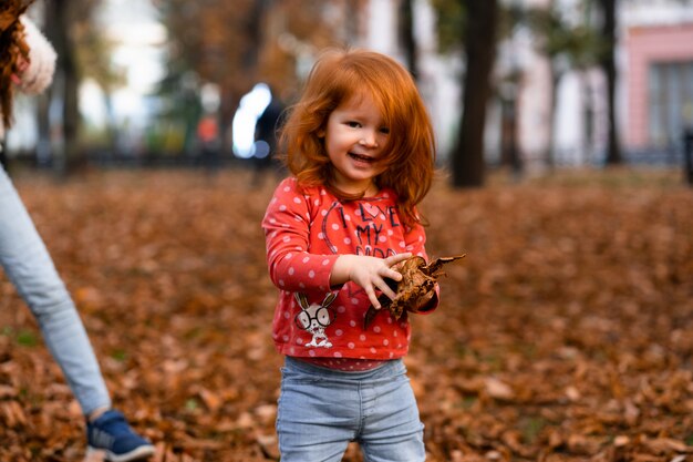 Closeup portret van schattige schattige lachende kleine roodharige kaukasische meisje kind spelen met droge bladeren staan in de herfst herfst park buiten, in de camera kijken, gelukkige levensstijl jeugd concept