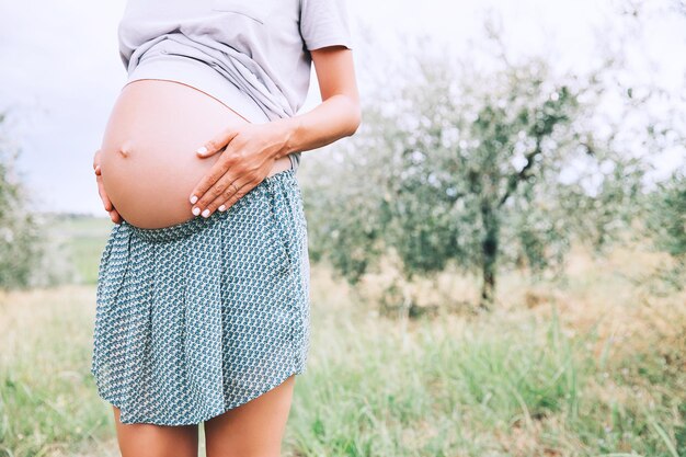 Closeup of pregnant belly in nature outdoors copy space Pregnant woman holds hands on belly on natural background of green grass Pregnancy expectation birth of new life concepts