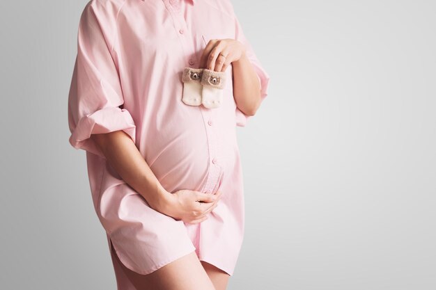 Closeup pregnant belly with cute tiny baby socks Concept of pregnancy gynecology preparation Beautiful pregnant woman in pink shirt dress in white background Young mother waiting for baby birth