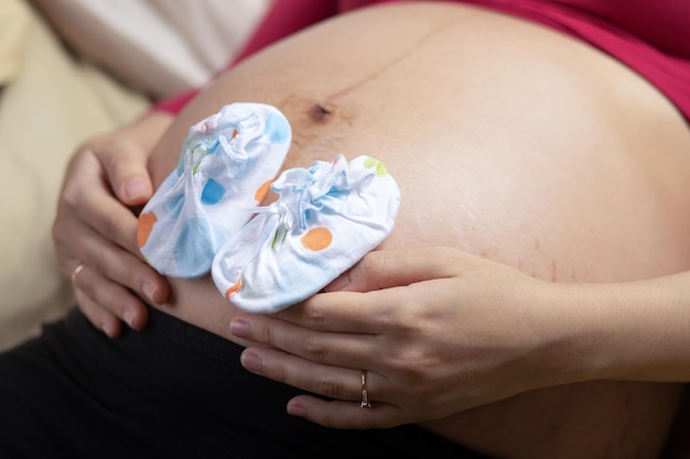 Крупный план беременной женщины на большом животе с маленькой детской обувью Концепция беременности и ожидания ребенка