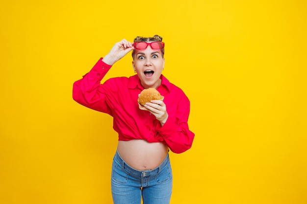 明るいピンクのシャツとジャンク フードを着た妊婦のクローズ アップ ハンバーガーと妊娠 不健康な食べ物を食べる妊婦のコンセプト 陽気な妊婦がファーストフードを食べる