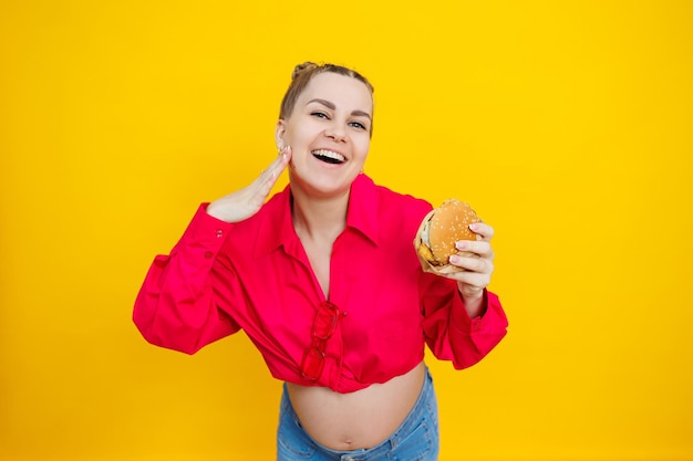 Closeup of a pregnant woman in a bright pink shirt and junk food Hamburger and pregnancy The concept of a pregnant woman eating unhealthy food Cheerful pregnant woman eats fast food