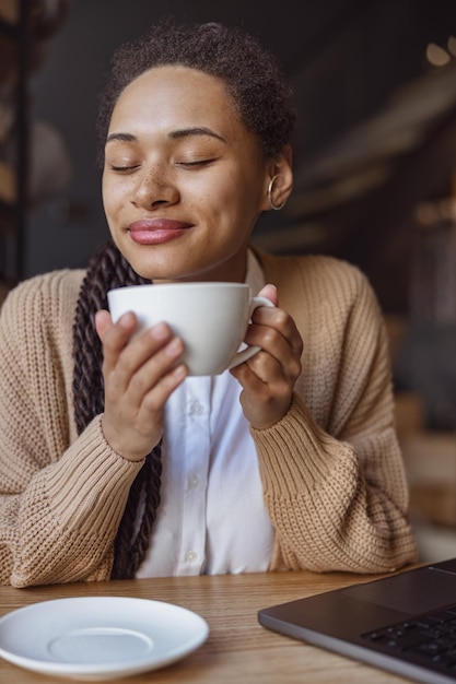 Closeup pretty ethnic woman holding cup with hot drink enjoying the aroma of cappuccino while chilling in coffee shop