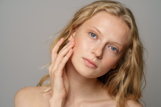 Primo piano del viso di donna abbastanza giovane con gli occhi azzurri, capelli biondi ricci naturali, non ha trucco, tocca la sua pelle morbida, in piedi a torso nudo con le spalle nude, guardando la fotocamera. sfondo grigio studio