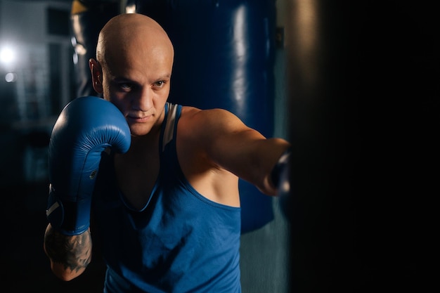 Closeup of professional bald boxer male training punches\
beating punching bag in sport club with dark interior