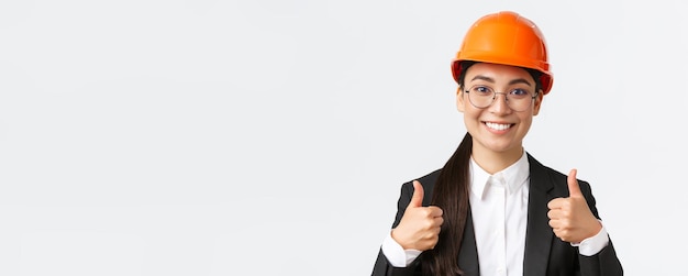 Closeup of professional pleased asian female chief engineer architect in business suit and safety helmet showing thumbsup in approval give permission recommend construction company