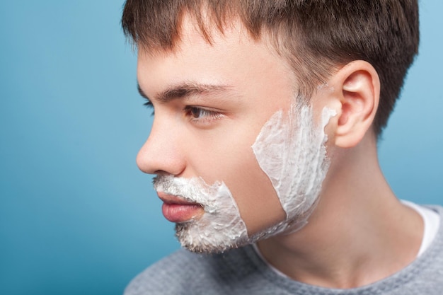 Closeup profile of young confident man looking aside with shaving foam on face showing smooth soft skin on shaved part of cheek moisturizing cosmetics indoor studio shot isolated on blue background