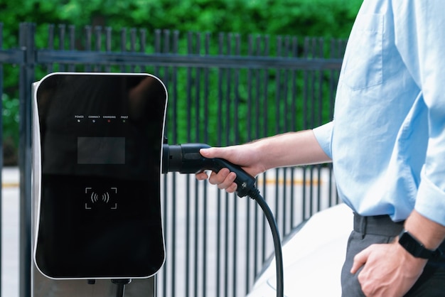 クローズ アップの進歩的なビジネスマンは、充電ステーションから EV に充電器のプラグを差し込みます