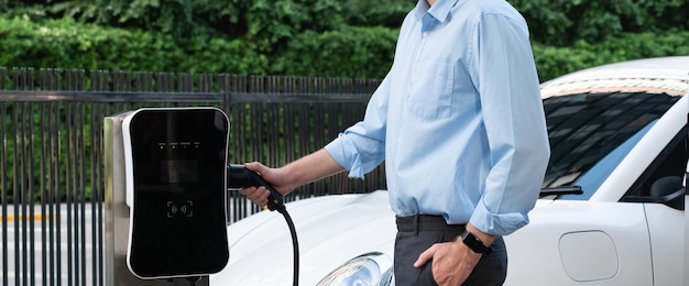 クローズ アップの進歩的なビジネスマンは、充電ステーションから EV に充電器のプラグを差し込みます