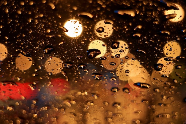 Foto gocce di pioggia in primo piano sulla superficie del parabrezza dell'auto, con luci stradali urbane sfocate di notte